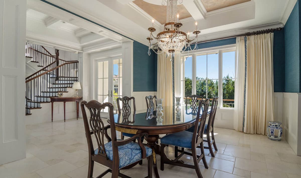 Dining room with round table, chairs, chandelier, large windows, and a curved staircase.