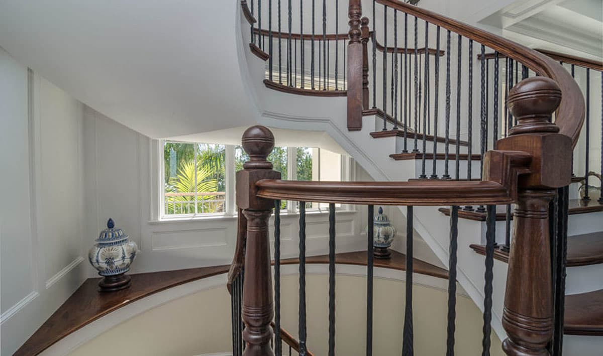 Interior view of a curved staircase with dark wood banisters and black spindles. Windows show greenery outside.