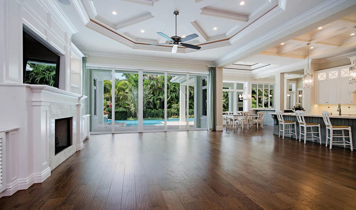 Great room interior with fireplace, coffered ceiling, fan, and adjacent kitchen with island. Large sliding glass doors open to outdoor pool area.