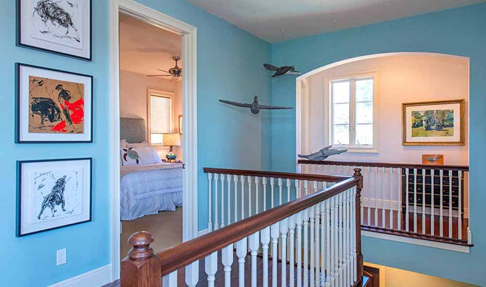 Interior hallway with light blue walls, white balusters, wood railings, framed art, and view into a bedroom.