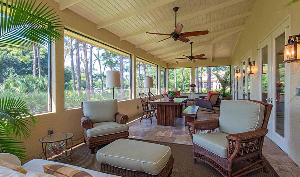 Covered porch with exposed beam ceiling, ceiling fans, and large windows overlooking a wooded area.