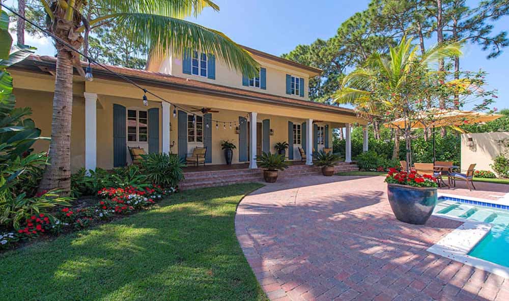 House plan exterior. Two-story with covered porch, shutters, and columns. Includes landscaping with palm trees and a brick paver driveway.