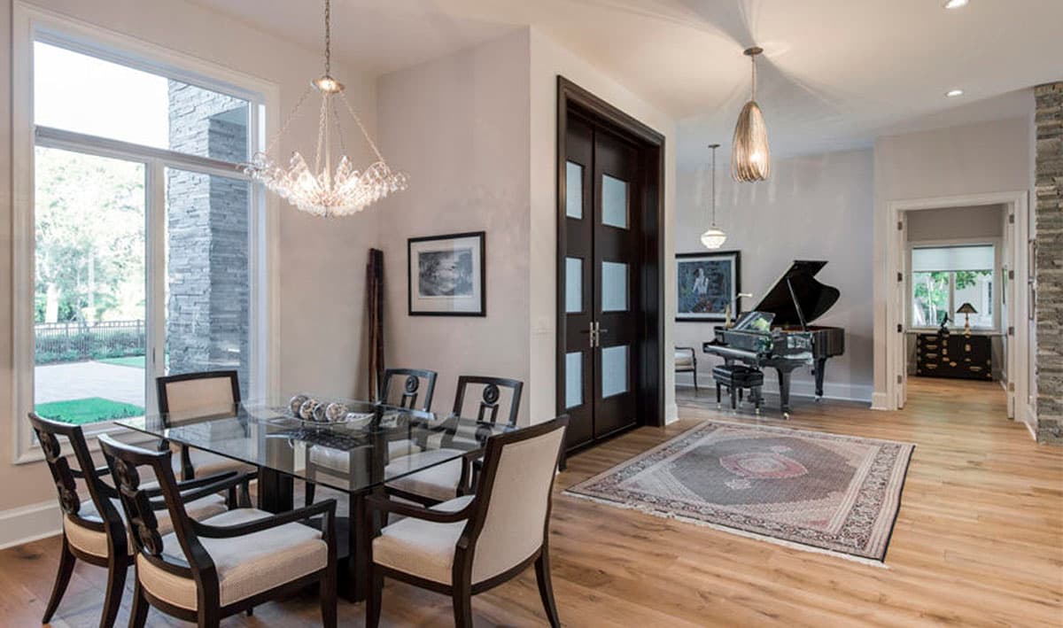 Modern dining room with glass table and grand piano in adjacent living area. Features stone exterior accents.