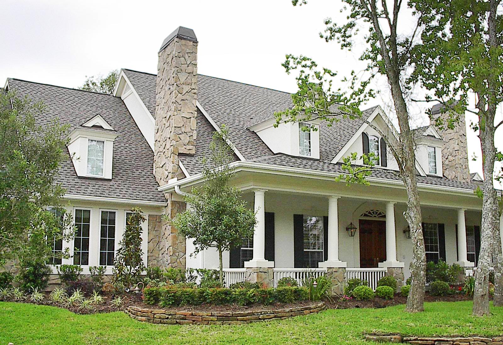 House plan exterior: Two-story home with stone chimney, prominent covered porch supported by columns, and dormer windows.
