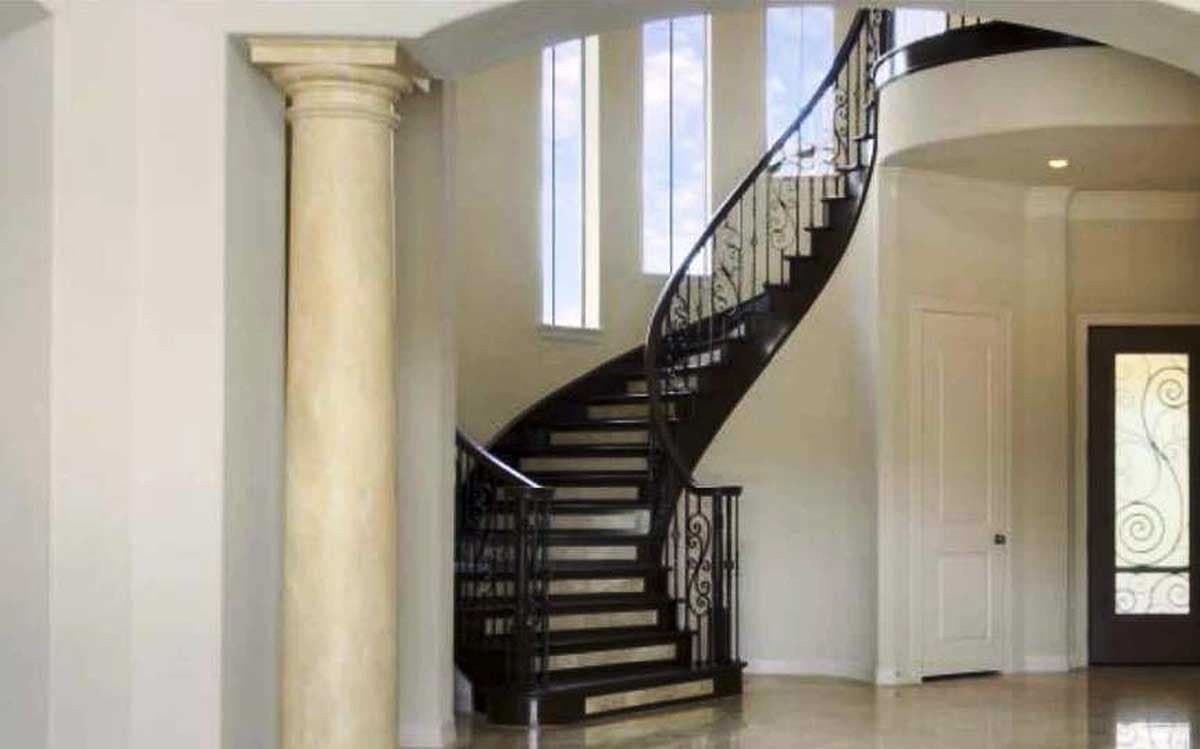 Interior view of a house with a curved staircase, ornate metal railing, classical column, and tall windows.