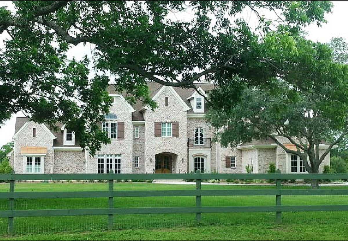 House plan exterior. Two-story brick home with gabled roof, multiple dormers, arched entry, and balcony. Includes a green fence.