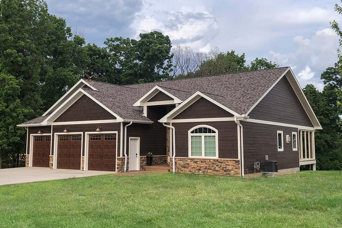 House plan exterior. Two-story with three-car garage, gable roof, and stone veneer accents. Features include an arched window and covered porch.