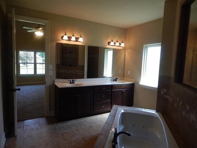 Primary bathroom with double vanity, jetted tub, and doorway to bedroom with ceiling fan and large window.