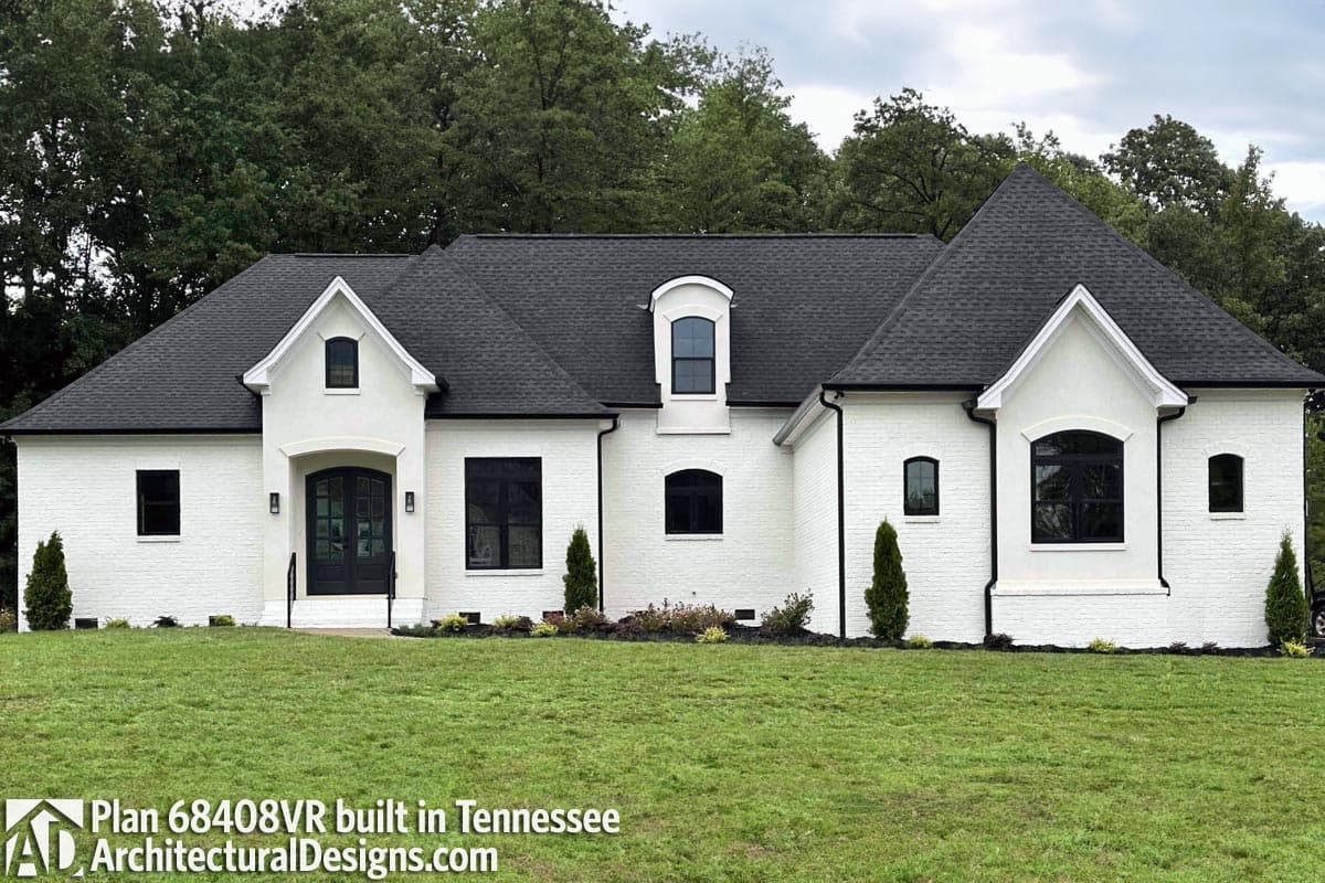 House plan exterior: White brick home with a prominent gable, arched dormer, and dark-trimmed windows.