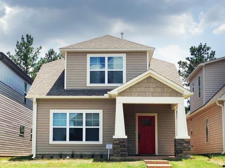 House plan exterior. Two-story home with a gabled roof, a front porch with columns, and a dormer. Vertical siding and a red front door are visible.