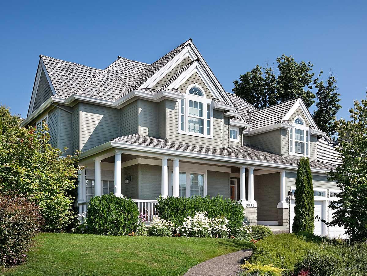 Two-story house plan exterior with wide covered porch, varied gables, arched dormer windows, and shingle roof.