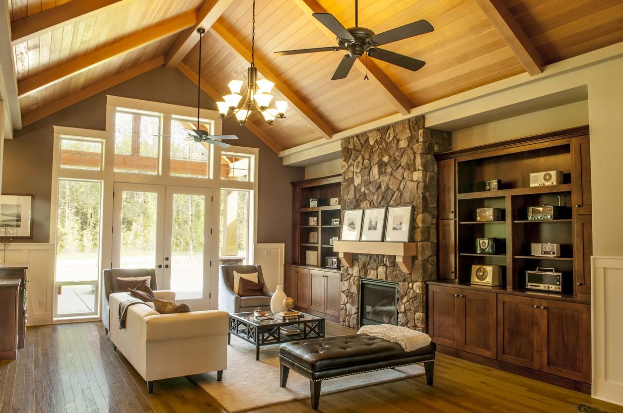 Living room interior with vaulted wood-paneled ceiling, stone fireplace, built-in cabinetry, and large windows.