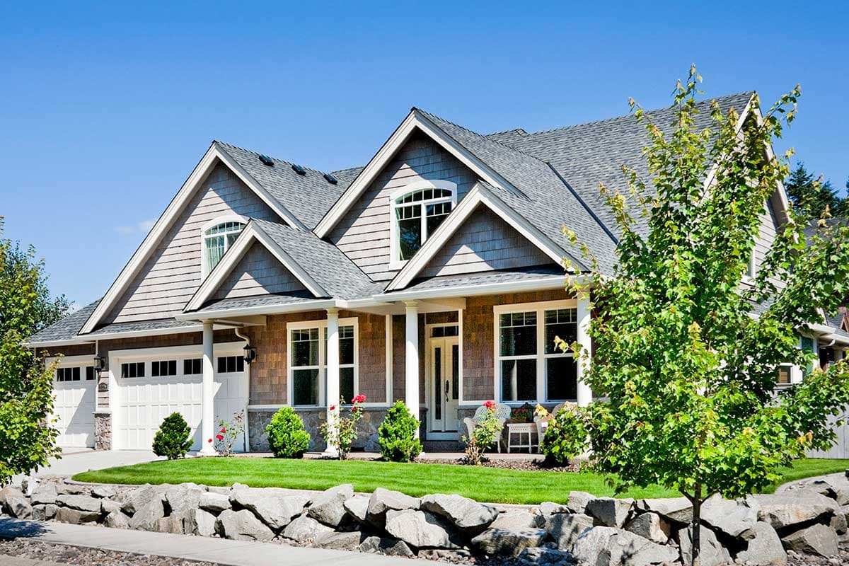 House plan exterior. Craftsman style, two-story house with gables, a covered porch, and a side-entry garage. Features shake siding and a stone wall.