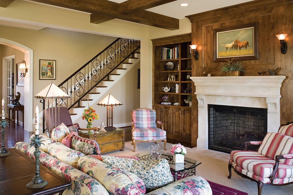 Interior view of a living room with exposed wood beams, a stone fireplace, built-in bookshelves, and a staircase with ornate metal railing.