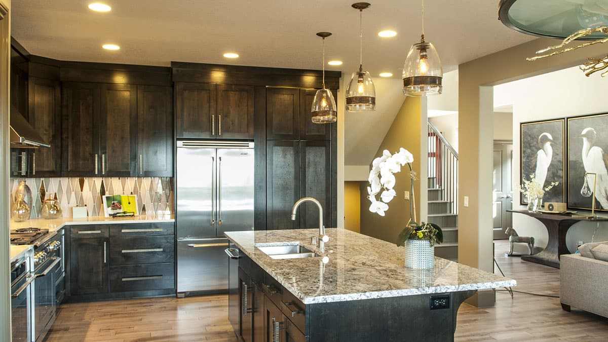 Kitchen with dark wood cabinets, granite island with sink and faucet, stainless steel refrigerator, and patterned backsplash. Open to living area.