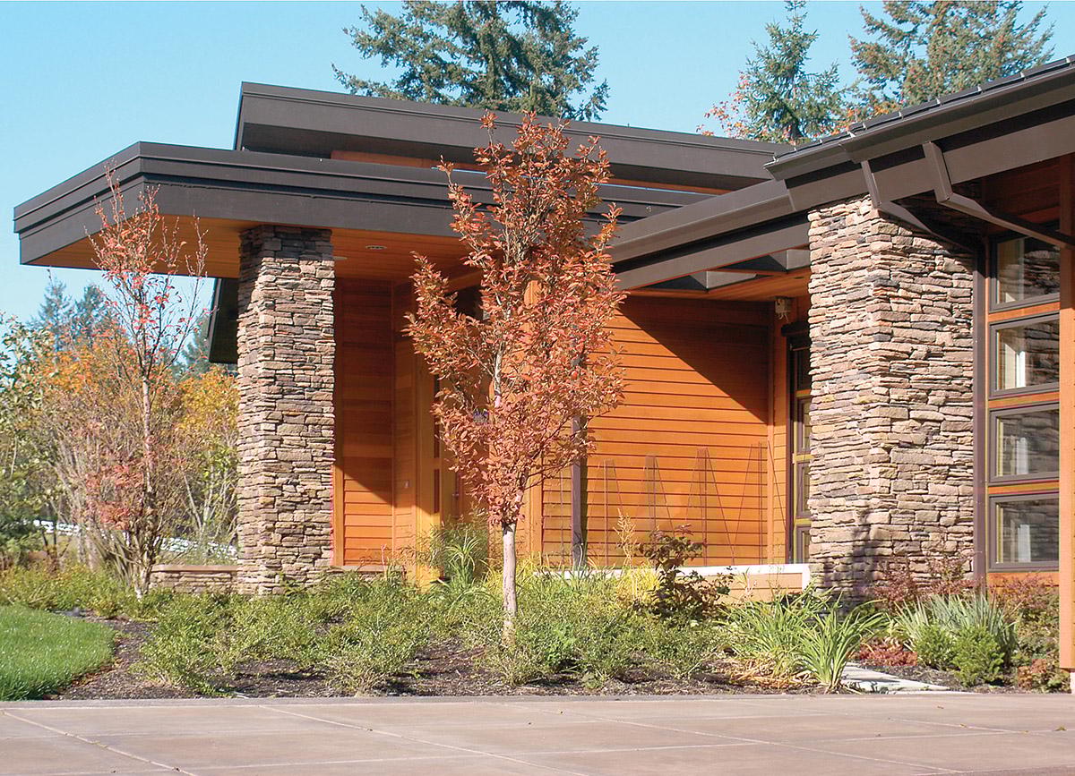 Modern house exterior with stacked stone columns, wood siding, layered rooflines, and large windows.