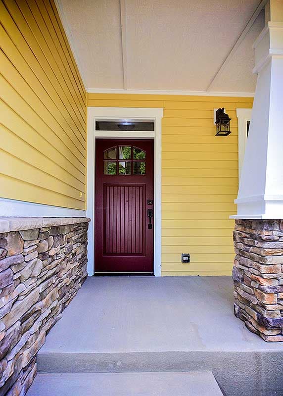 House plan exterior: Craftsman style, yellow horizontal siding, stone foundation, covered porch with paneled door and arched window.