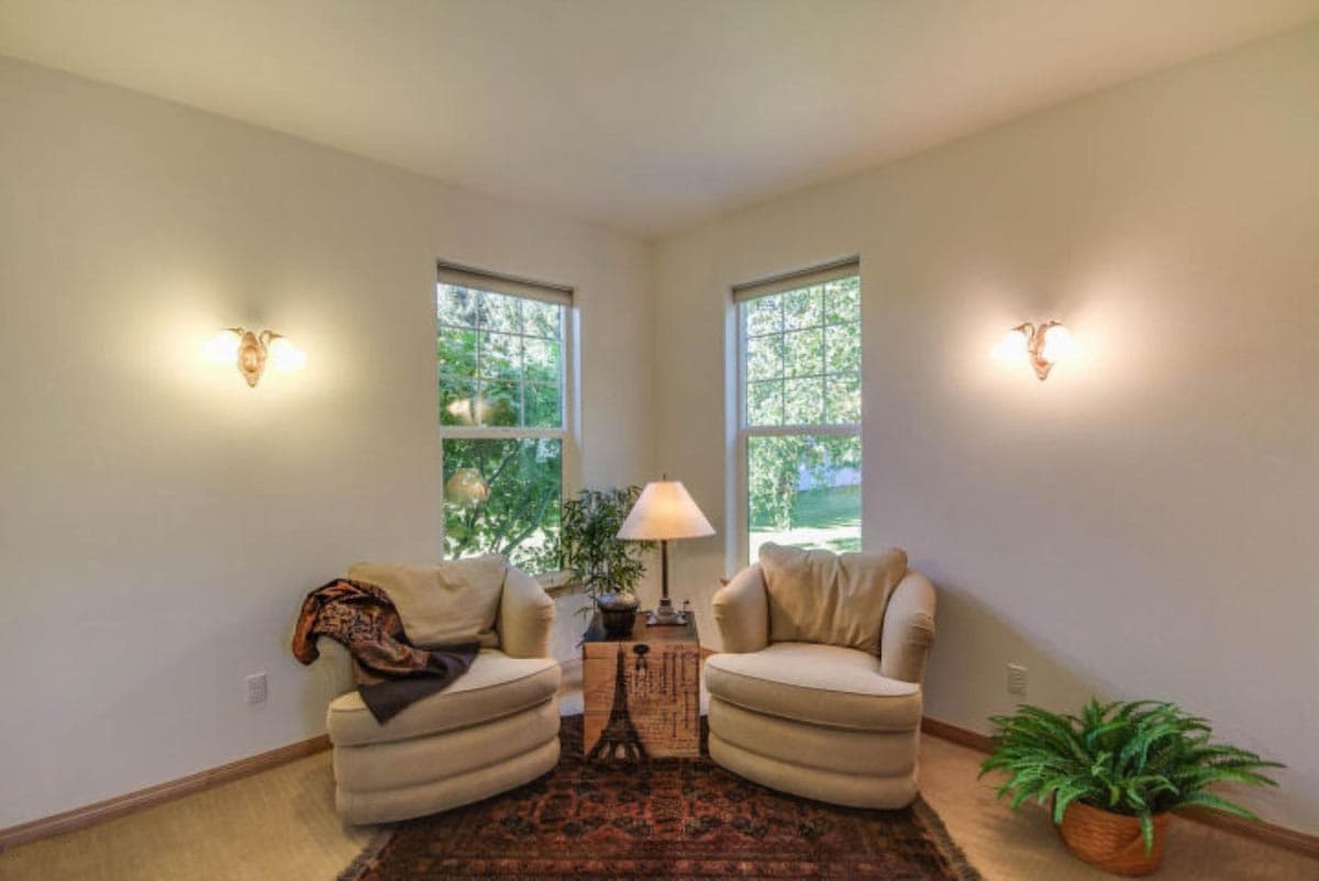 Two upholstered chairs flank a table with a lamp between two windows. Wall sconces illuminate the room.