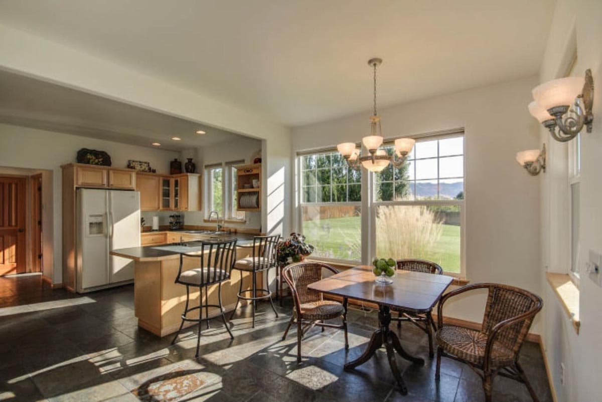 Kitchen and dining area with island, bar stools, wood table, chairs, and large windows overlooking backyard.