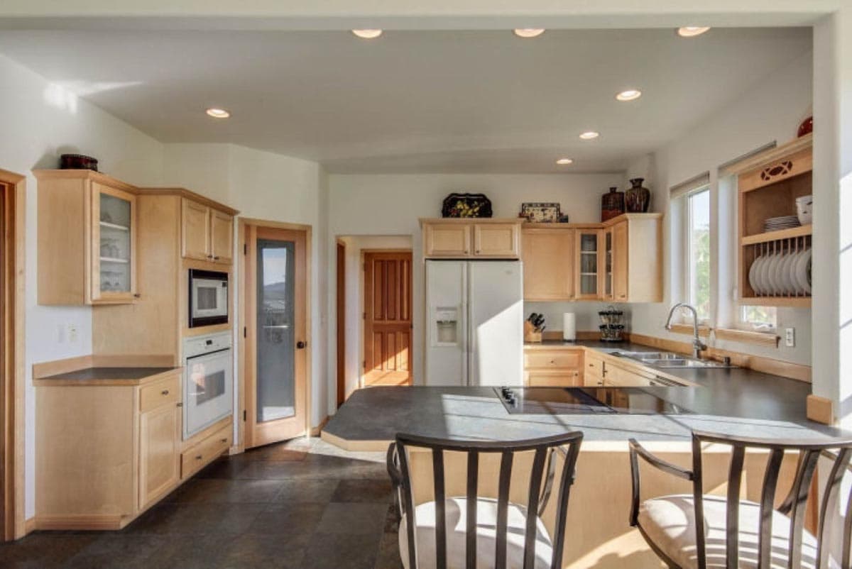 Kitchen with light wood cabinetry, dark countertops, island cooktop, stainless steel refrigerator, and built-in oven.
