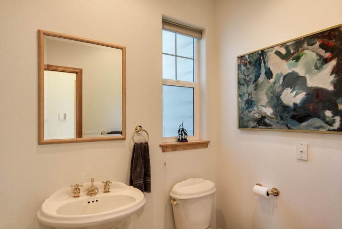Powder room interior with pedestal sink, toilet, framed mirror, and abstract artwork above toilet paper holder.