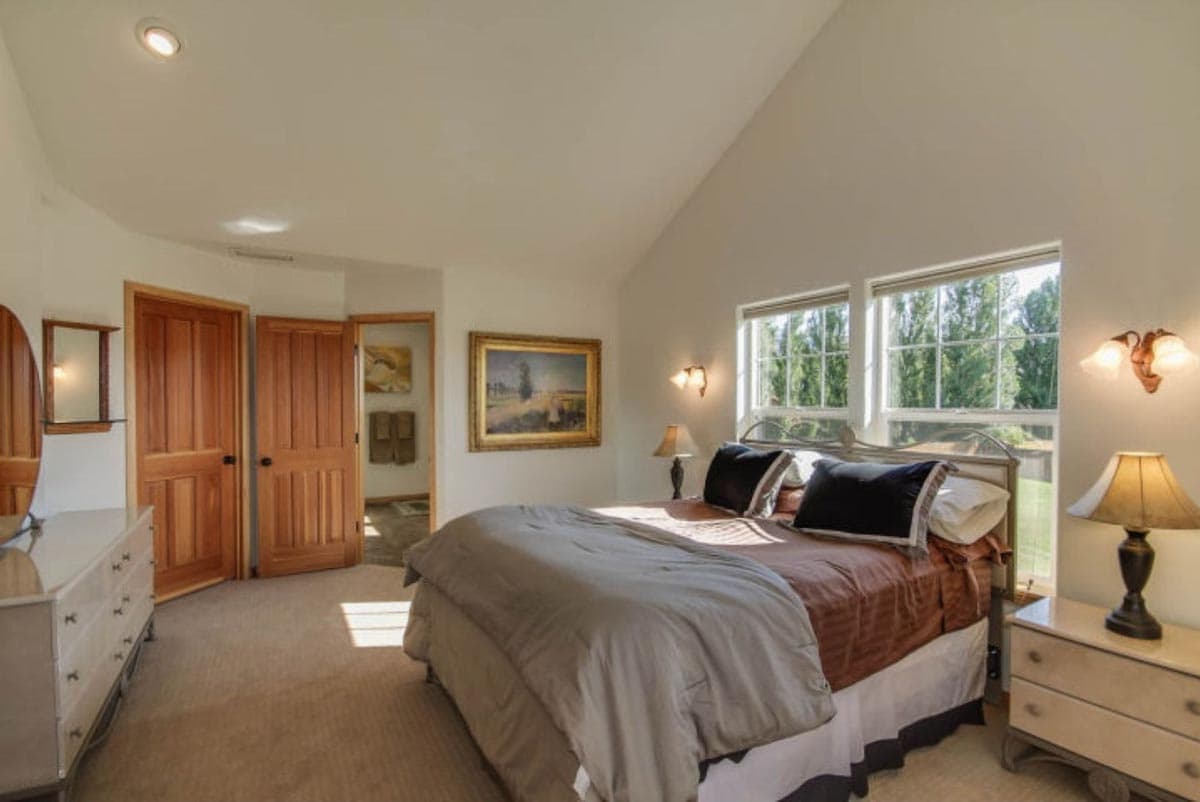 Bedroom with vaulted ceiling, double doors, and two windows overlooking trees.