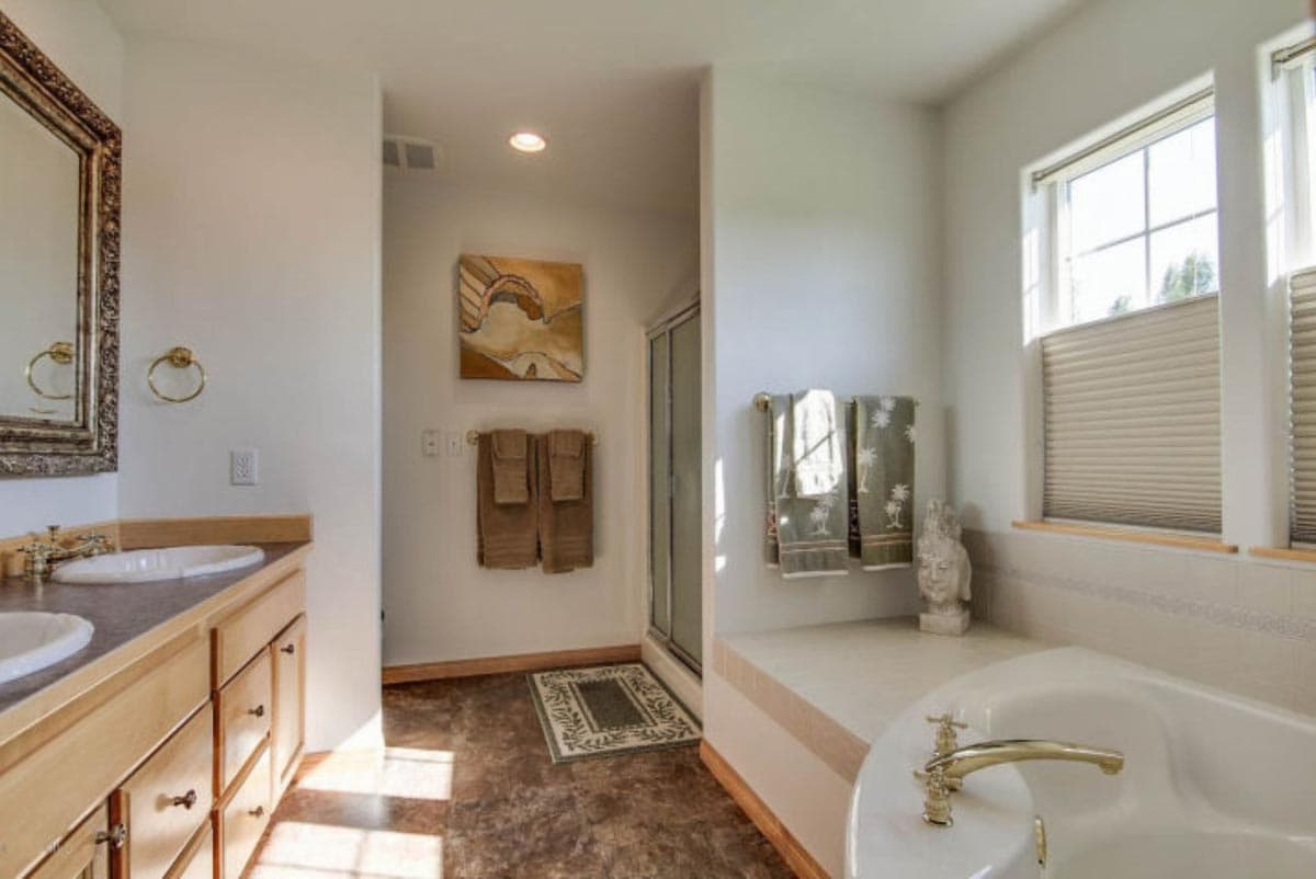 Bathroom featuring double vanity, glass-enclosed shower, soaking tub, and window with blinds.