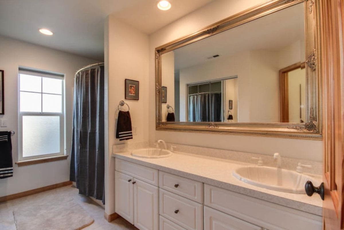 Bathroom with double vanity, large ornate mirror, and walk-in shower with dark curtain. Window with grid pattern.