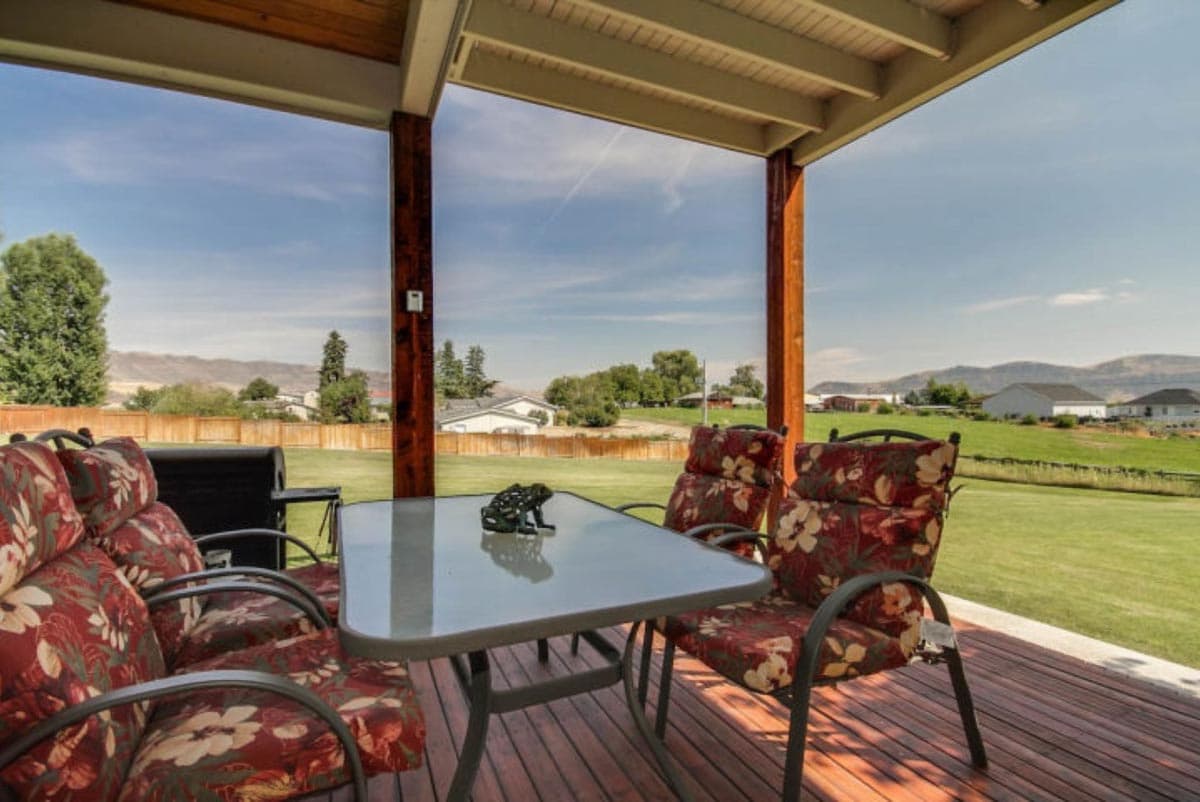 Covered porch with seating and table overlooks a grassy yard with distant homes and hills.