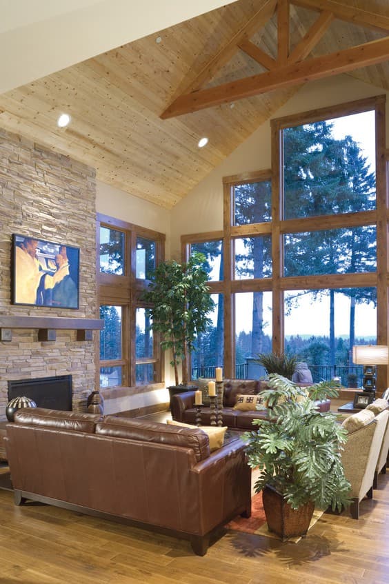 Great room with stone fireplace, wood-paneled vaulted ceiling with exposed beams, and tall windows overlooking forest.