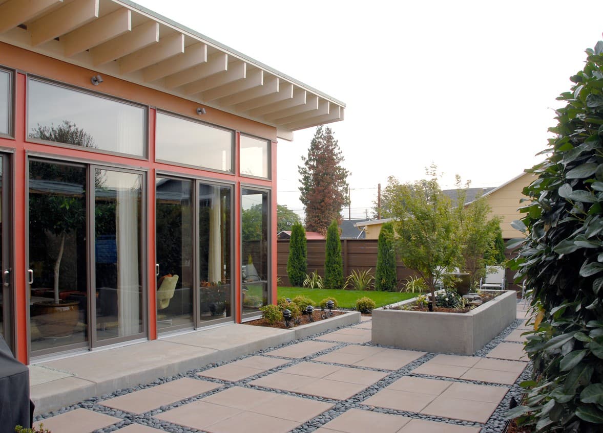 Mid-century modern house exterior with large glass windows, exposed rafters, and a patio with square pavers and river stones.