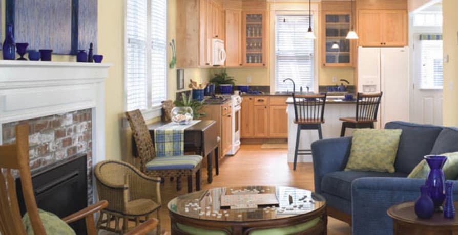 Interior view of a living area with a fireplace, dining nook, and open kitchen with light wood cabinets and a breakfast bar.