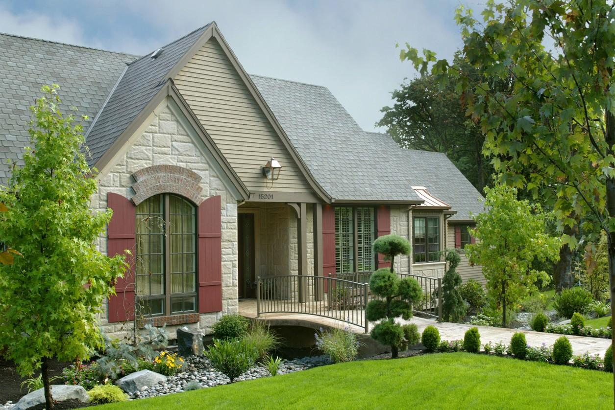 House plan exterior with stone and vinyl siding, arched window, red shutters, covered entry with bridge over a water feature.