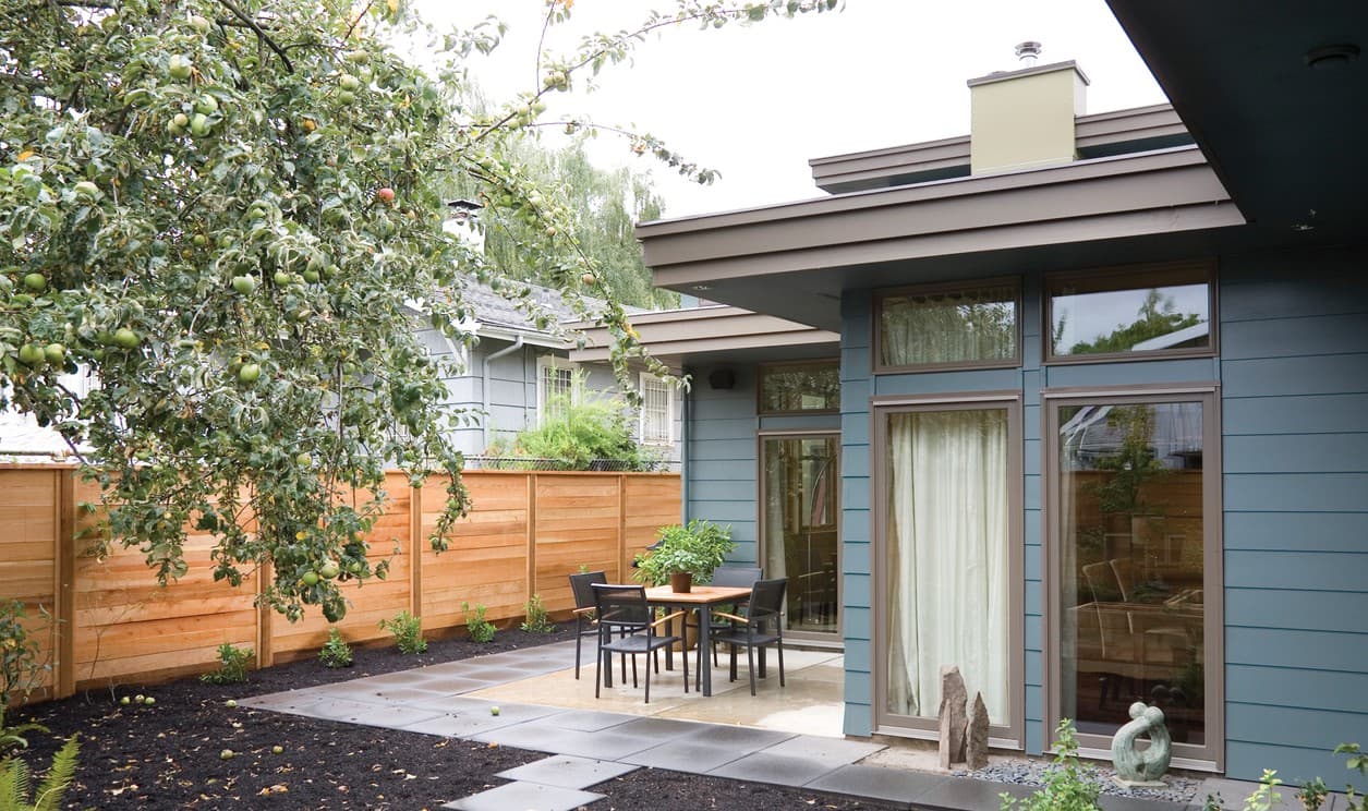 Modern house exterior with horizontal siding, large windows, overhanging roof, and a patio with outdoor dining.
