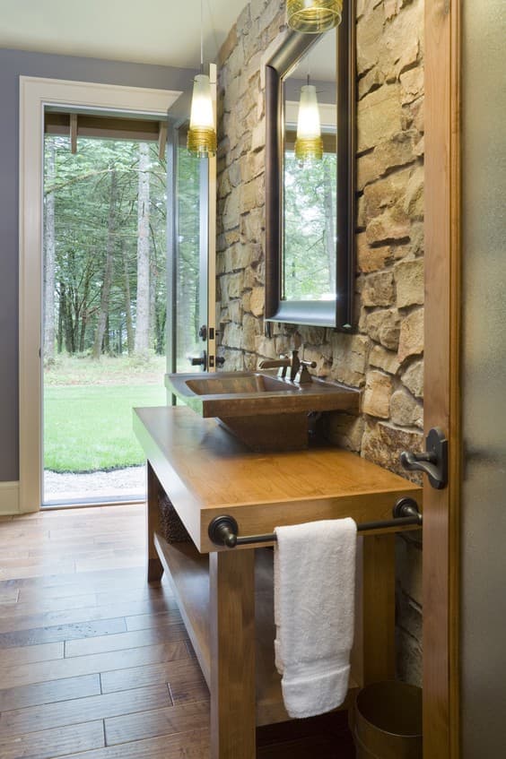 Bathroom interior with stone accent wall, wood vanity, stone vessel sink, and glass door opening to a wooded backyard.