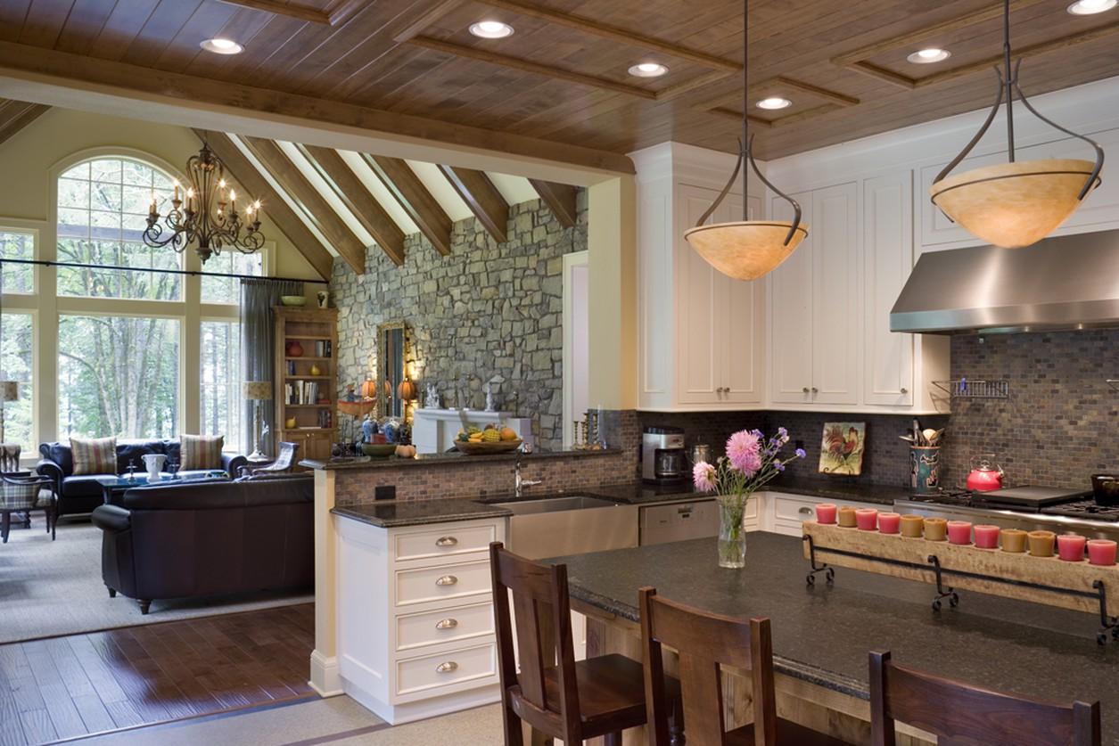 Open-concept living area with wood-beamed vaulted ceiling, stone accent wall, and kitchen with island, pendant lights, and stainless steel appliances.