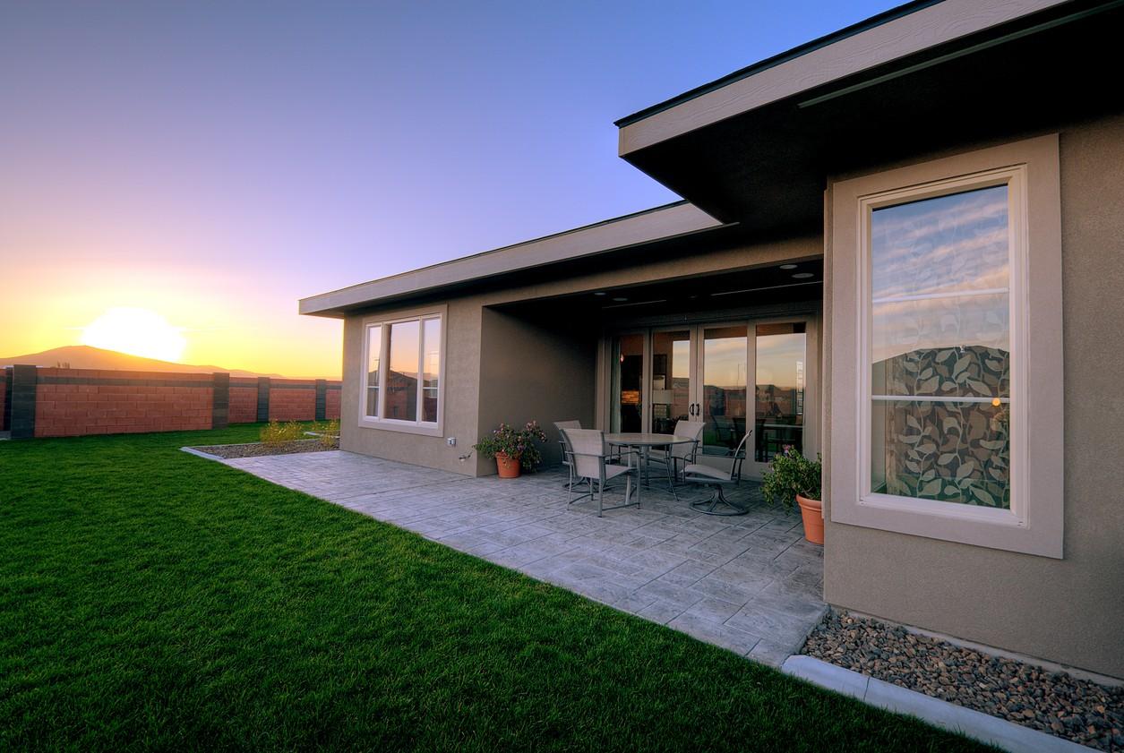 Modern house exterior with a covered patio, large sliding glass doors, and multiple windows. A concrete patio extends to a lawn.