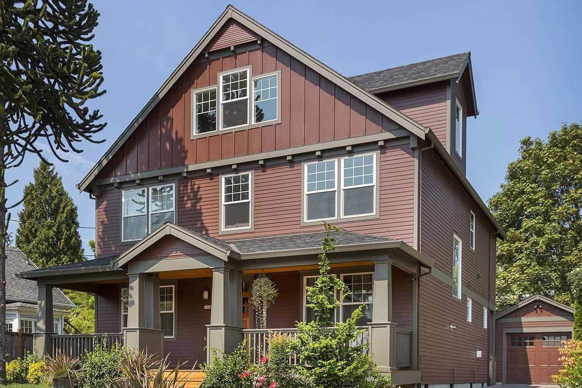 House plan exterior: three-story, dark red siding, prominent gables, deep front porch with square columns, and a detached two-car garage.