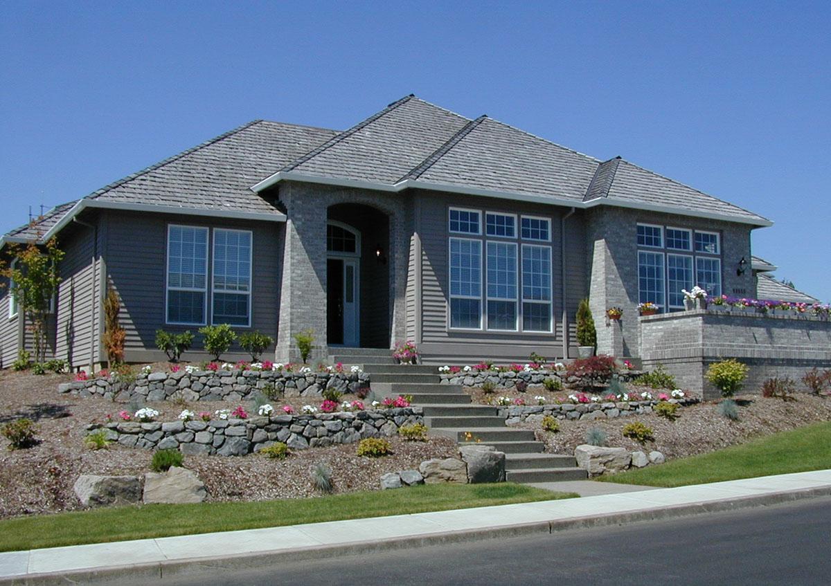 House plan exterior: One-story home with arched entryway, large windows, shingle roof, and tiered stone landscaping.
