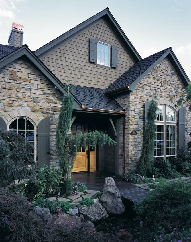 House plan exterior featuring stone and shingle siding, gabled dormer, arched windows, and wood garage doors.