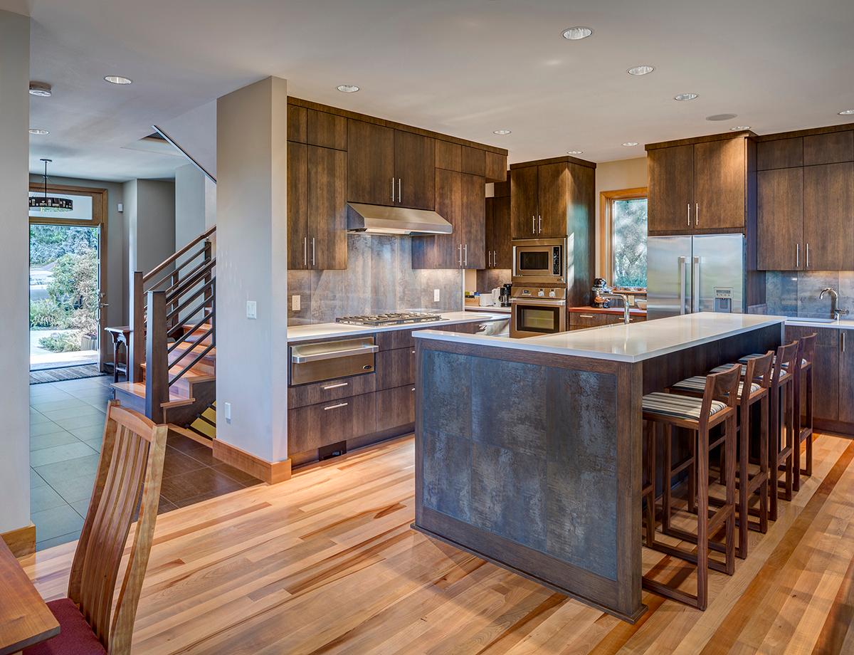 Modern kitchen with dark wood cabinets, quartz island with bar stools, stainless steel appliances, and open staircase.