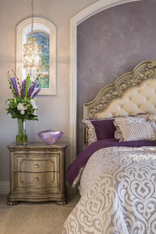Bedroom interior with ornate headboard, tufted upholstery, patterned bedding, decorative nightstand, and arched window with chandelier.