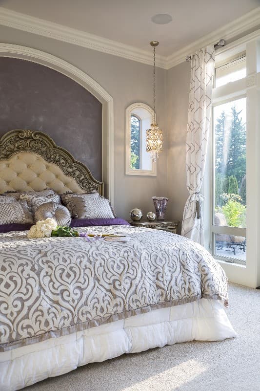 Bedroom interior with tufted headboard, ornate bed linens, decorative pillows, and large window with patterned curtains.