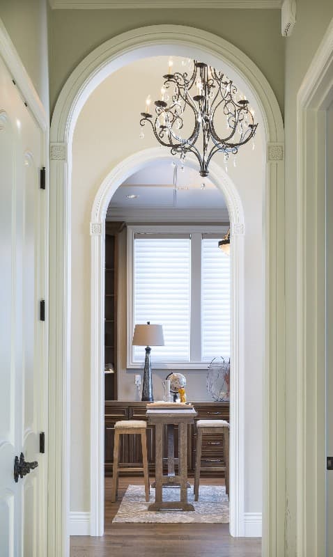 Interior view looking into a room with a decorative chandelier, two bar stools, and a tall table in front of a window with blinds.