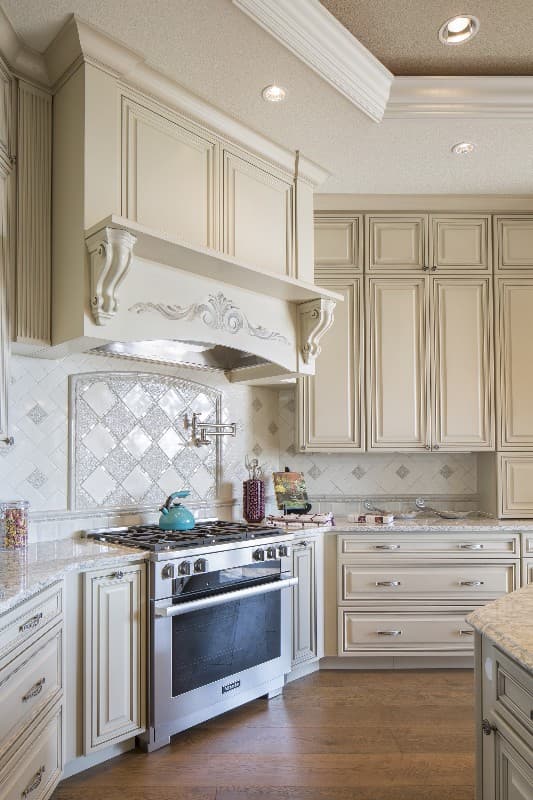 Kitchen interior with stainless steel range, ornate hood, cream cabinetry, and patterned tile backsplash.
