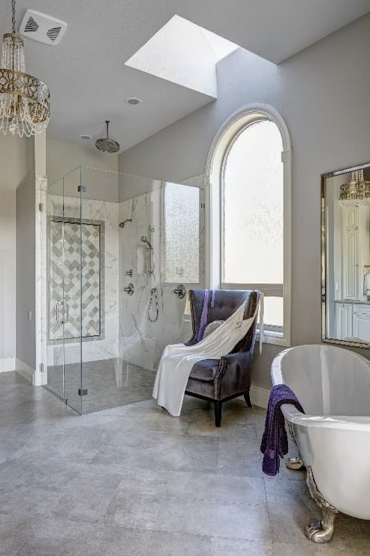 Spacious bathroom interior with glass-enclosed shower, freestanding tub, arched window, skylight, and chandelier.