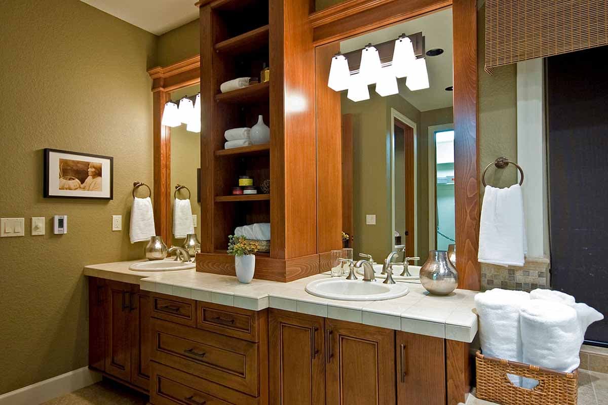 Bathroom vanity with double sinks, wood cabinetry, built-in shelving, and tiled countertop.