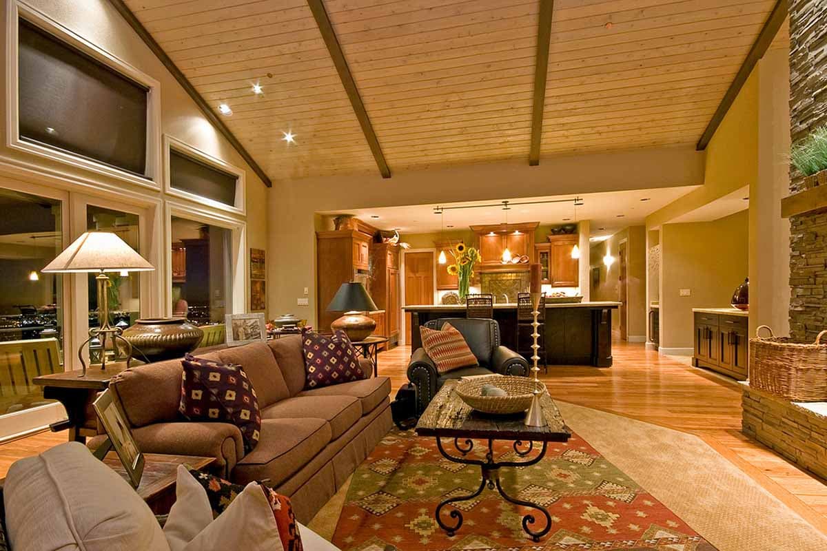 Great room interior with vaulted wood ceiling, stone fireplace, large windows, and open kitchen with island.