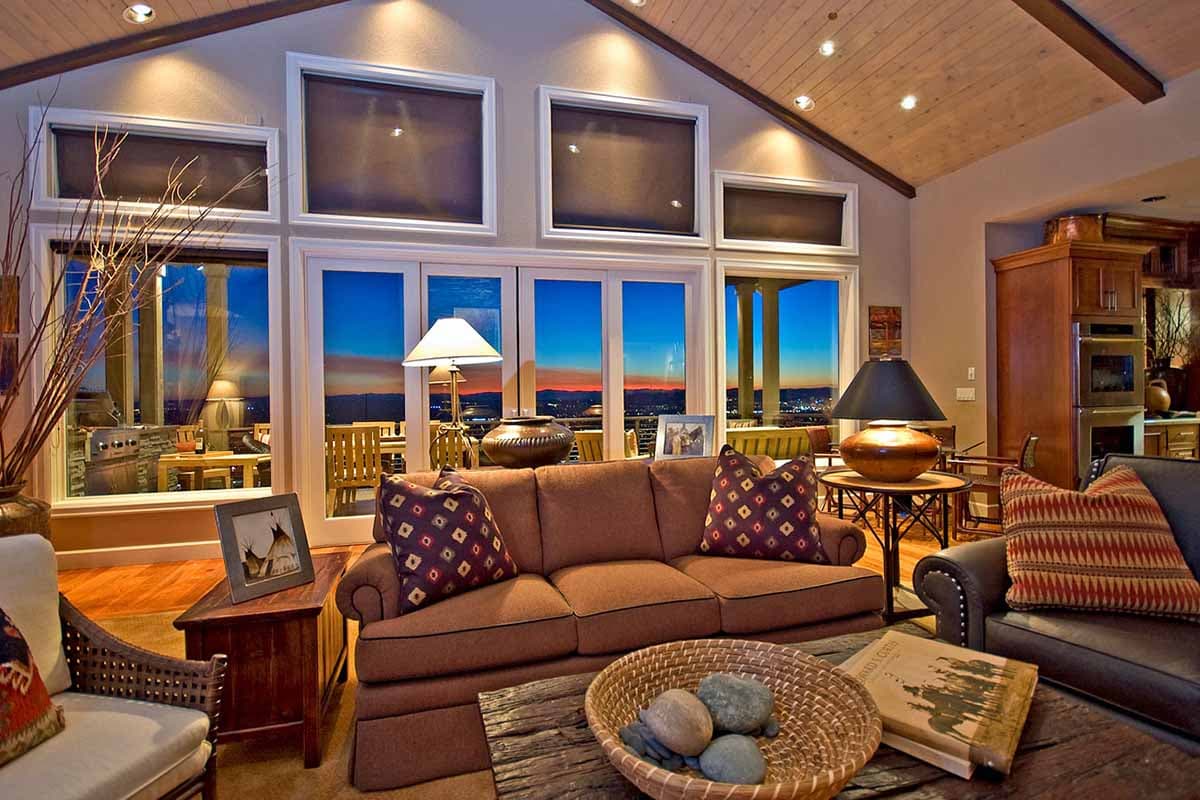 Interior view of a living room with vaulted wood ceiling, large windows overlooking a sunset, and a rustic furniture arrangement.