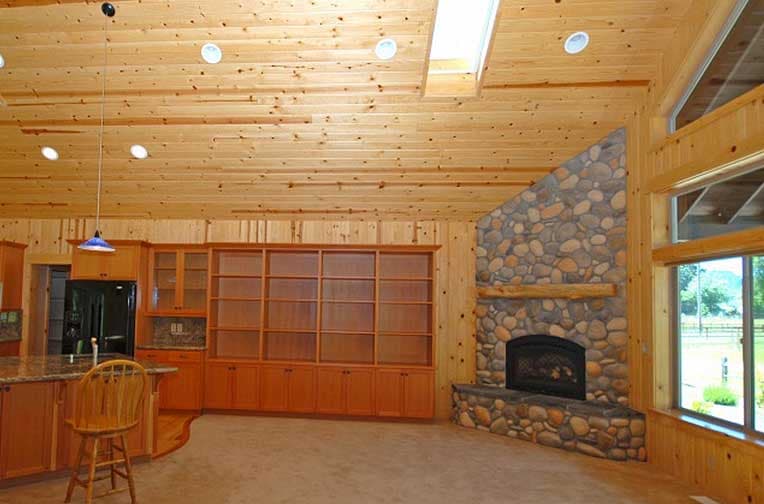 Interior view of open-concept living space with stone fireplace, built-in shelving, and wood plank ceiling with skylight.